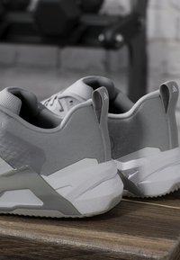 Paire de chaussures de sport grises et blanches posées sur une surface en bois, montrant les détails à l'arrière et sur le côté avec des languettes de traction marquées.