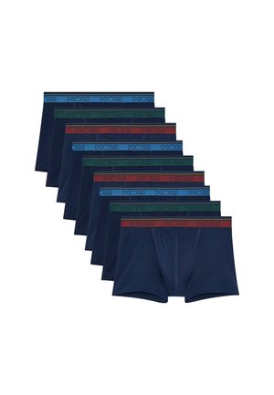 Muskelöser Mann steht barfuß in dunkelblauen Boxershorts mit rotem Bund vor einem einfachen hellen Hintergrund.