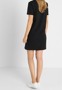 Černé šaty s krátkým rukávem, rovného střihu, délka ke kolenům, vyrobené z měkké tkaniny, spárované s bílými teniskami.