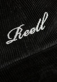 Black corduroy cap with white embroidered "Reell" logo. Features vertical ribbing for texture. Rounded peak and structured design.