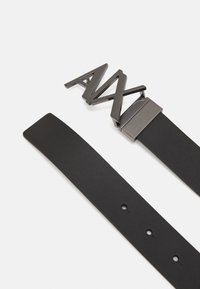 Sort læderbælte med en tekstureret overflade, prydet med en iøjnefaldende metalspænde i et geometrisk design med skarpe vinkler. Flad rem med huller til justering.