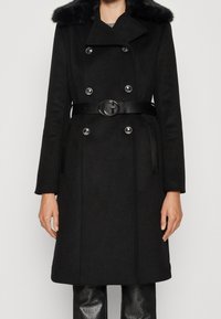 Zwart dubbelbreasted jasje met zilveren knopen, bontkraag en zwarte riem met ronde metalen gesp, gedragen over zwarte glanzende broeken.