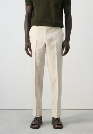 Personne à la peau foncée portant un pantalon crème sur mesure, une chemise vert olive foncé et des sandales en cuir marron foncé, debout sur un sol gris.