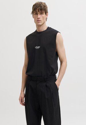 Νεαρός άνδρας με ανοιχτό καστανά μαλλιά, φορώντας αμάνικο μαύρο τοπ με λευκό λογότυπο και μαύρο παντελόνι με λεπτές ρίγες, στέκεται μπροστά σε λευκό φόντο.