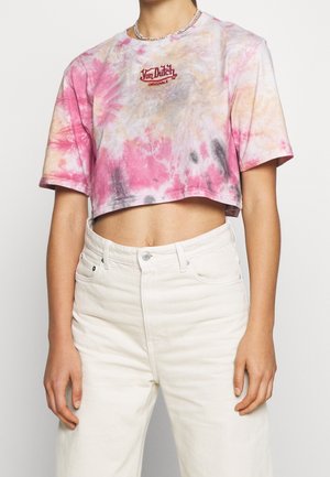 Kvinde iført cropped T-shirt i pink, orange og grå tie-dye med "Von Dutch Originals"-logo og højttaljede off-white bukser, hænderne afslappede.