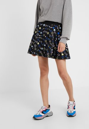 Personne portant une jupe noire avec des motifs d’étoiles bleues, jaunes et blanches, un haut gris à manches longues et des baskets blanches, bleues, roses et noires.