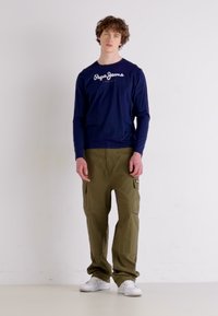 Tengerészeti kék hosszú ujjú póló "Pepe Jeans" logóval, olajzöld cargo nadrággal, oldalzsebekkel párosítva; a megjelenést fehér sportcipők egészítik ki.