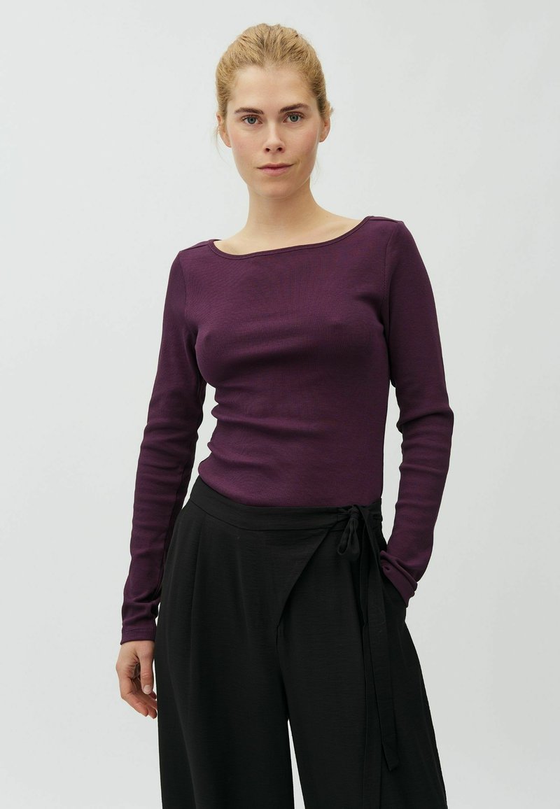 Femme à la peau claire et aux cheveux blonds attachés, portant un haut violet à manches longues et un pantalon noir ample, debout devant un fond clair uni.