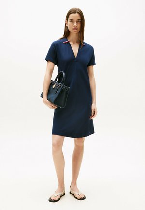 Marine jurk met een V-hals en korte mouwen, voorzien van een rode accent bij de kraag. Zwarte handtas met leertextuur. Beige slippers.