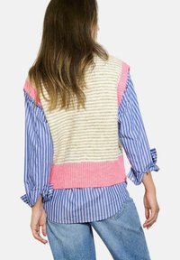 Bezramenný pletený svetr v béžových a růžových pruzích, nošený přes modro-bíle pruhovanou košili. Výrazný žebrovaný lem a jemná textura.