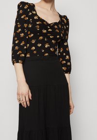 Kvinna i svart blommig puffärmad topp med rynkad halsringning och hög midja, svart lager på lager kjol, ringar synliga på vänster hand.