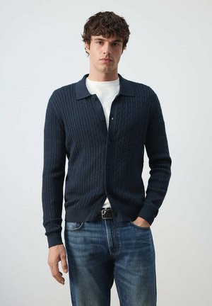 Hombre con un cárdigan azul marino de punto trenzado sobre una camisa blanca y vaqueros azules, de pie con una mano en el bolsillo.