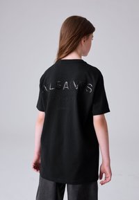 Fiatal személy hosszú hajjal, fekete, túlméretezett pólót visel, amelynek hátoldalán az "ALLSAINTS" felirat és más szövegek találhatók, egyszerű háttér előtt áll.