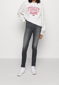 Jeans grigi con vestibilità slim, abbinati a una felpa grigia chiara con testo rosso "TOMMY JEANS" e sneakers bianche basse.