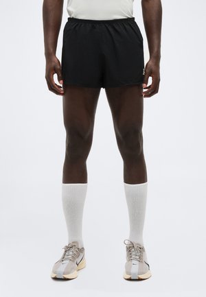 Moški v črnih športnih kratkih hlačah, belih nogavicah do kolen in bež tekaških copatih, stoji pred enakomerno svetlo ozadje.