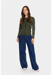 Roheline nööbiga kardigan, mis on valmistatud pehmest kudumikambris, on kombineeritud laia jalaga siniste teksapükstega. Mudel seisab, naeratades heledates kingades.