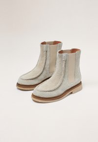 Bottines Chelsea beige avec tige en tissu texturé, panneaux latéraux élastiques et semelle en brun clair contrastant. Bout rond et détails de couture visibles.