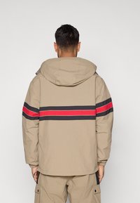 Κρέμα ζακέτα με κουκούλα, διαθέτει μαύρες και κόκκινες ρίγες στη πλάτη. Φτιαγμένη από ελαφρύ υλικό με ελαστικές μανσέτες.