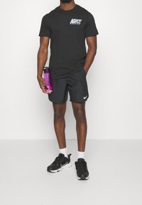 Černé tričko Nike s logem, sladěné s černými šortkami, bílými ponožkami a černými teniskami. Drží fialovou láhev na vodu.