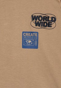 Beige t-skjorte med blå brodert "WORLD WIDE" og en blå grafisk etikett med teksten "CREATE FUTURE" med illustrasjon av en globus.