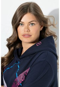 Felpa con cappuccio blu navy caratterizzata da vivaci motivi geometrici in rosa e blu, con un'etichetta tessuta che presenta un simbolo dell'infinito sul cappuccio.