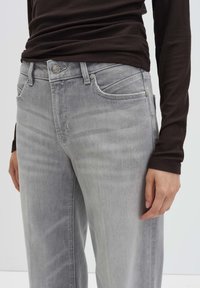 Close-up van een persoon in lichtgrijze jeans en een donker, langemouwen shirt, waarbij de zakken en knoopdetails van de jeans zichtbaar zijn.