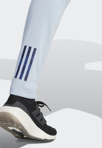 Lyseblå træningsbukser med tre marineblå striber ved anklerne. Båret med sorte sneakers, der har en struktureret overdel og en hvid sål.