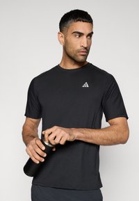 Μαύρο αθλητικό μπλουζάκι με κοντά μανίκια, στρογγυλή λαιμόκοψη και λευκό λογότυπο σε σχήμα τρίγωνο στο αριστερό στήθος. Το μοντέλο κρατάει ένα μαύρο παγούρι νερού.