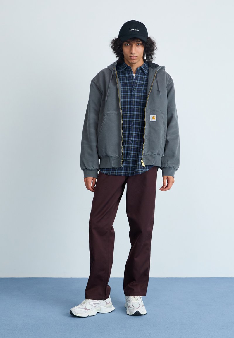 Grå zip-up hoodie med gul dragkedja, rutigt skjorta under, vinröda byxor och vita sneakers med grå detaljer, buren med en svart keps.