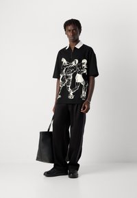 Man wearing black polo shirt with white boxing figures, black pants, and holding a black tote bag, standing against a plain white background.