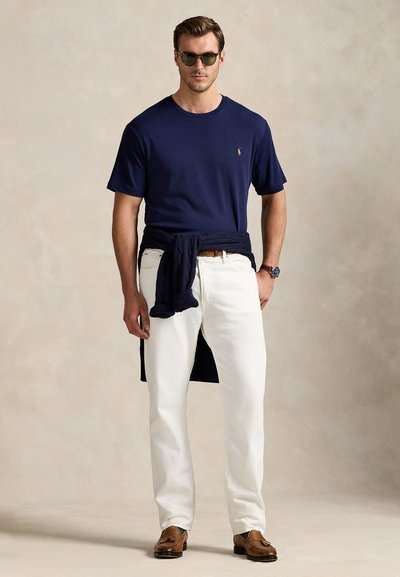 Bărbat purtând un tricou albastru închis, pantaloni albi, mocasini maro, ochelari de soare și un pulover albastru închis legat în jurul taliei, pe un fundal neutru.