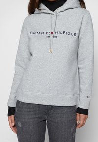 Sudadera gris con capucha, logo frontal que dice "TOMMY HILFIGER EST 1985" y puños negros acanalados. Tejido suave con un corte informal.