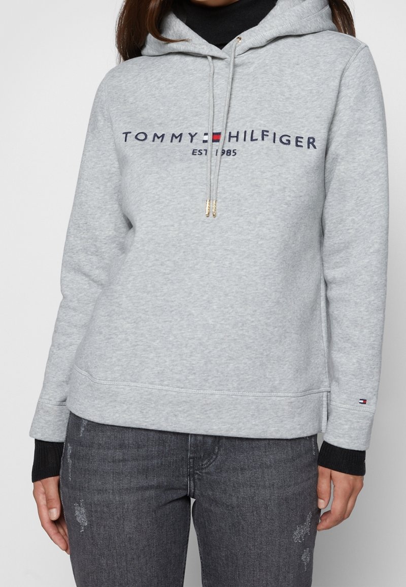 Sudadera gris con capucha, logo frontal que dice "TOMMY HILFIGER EST 1985" y puños negros acanalados. Tejido suave con un corte informal.
