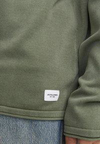 Zöld kötött pulóver texturált anyagból, alján fehér címkével, rajta a "JACK & JONES EST. 1990" felirattal és kerekített hem.
