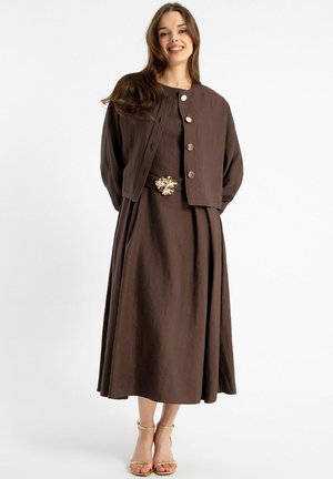 Uśmiechnięta kobieta w brązowej, zapinanej na guziki kurtce nałożonej na pasującą midi sukienkę z złotą klamrą paska i złotymi sandałami na obcasie z paskami.