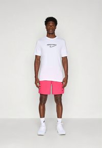 Camisola branca de manga curta Nike Pro com logótipo preto, combinada com calções cor de rosa e meias brancas com logótipo preto da Nike. Em pé sobre um fundo neutro.