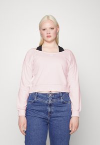 Camisola cropped rosa clara com mangas longas e decote redondo, combinada com jeans de ganga azul de cintura alta. Apresenta um detalhe de logótipo discreto.