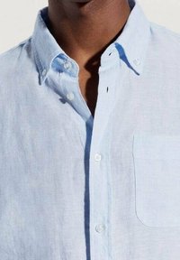 Camisa de lino azul claro con cuello abotonado, que cuenta con un bolsillo en el pecho y botones blancos, exhibiendo una tela suave y texturizada.