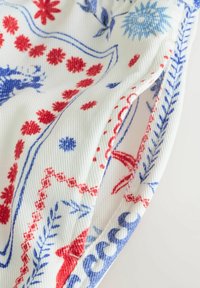 Tissu blanc avec des motifs floraux et géométriques bleus et rouges, présentant des plis et une texture visible du matériau tissé.