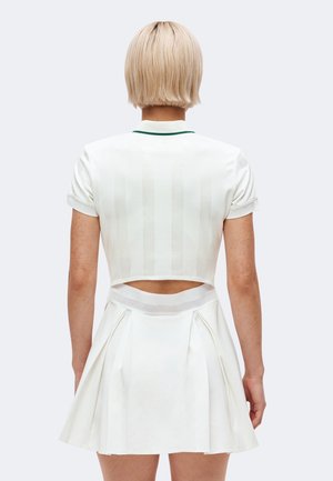 Personne aux cheveux blonds courts portant un haut court blanc et une jupe plissée avec une ceinture en mesh, vue de dos.