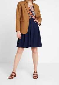 Blazer marrone sopra una maglia floreale, abbinato a una gonna blu navy e scarpe con tacco a strappo marroni. Le caratteristiche includono un taglio sartoriale e un design della gonna svasata.