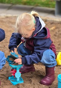 Barn som använder en blå sandleksak med rosa detaljer i en sandlåda. De har på sig en marinblå och vinröd jacka, blå byxor och vinröda stövlar.