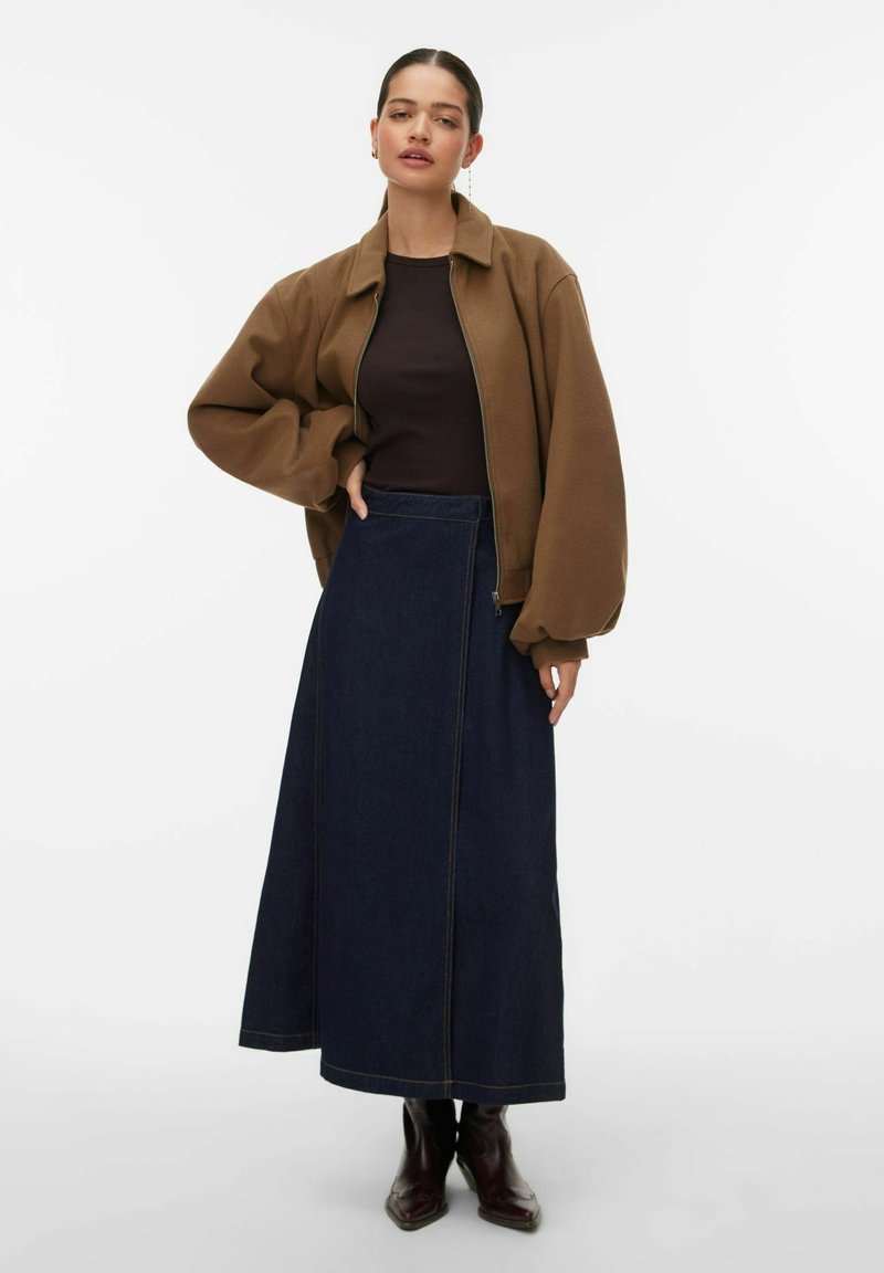 Veste bomber en polaire marron, haut noir ajusté, longue jupe en denim avec coutures contrastantes, et bottes en cuir marron. Palette de couleurs simple.