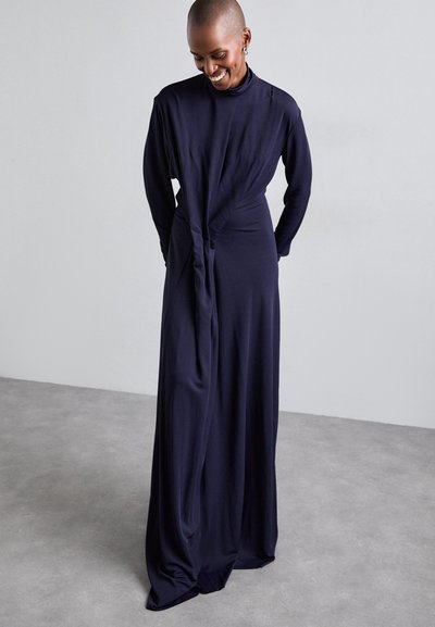 Navy long-sleeve dress with a high neckline, draped front, and twisting detail at the waist; made of soft, flowing fabric.