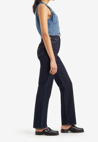 Femme debout de profil portant un gilet en jean sans manches, un jean bleu foncé taille haute et des chaussures noires à enfiler avec des boucles.