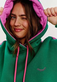 Zelená mikina s ružovým podšíváním a šnúrkami. Na prednej strane má jednoduchý vzor očných rias vyšívaný. Mäkká textúra látky.