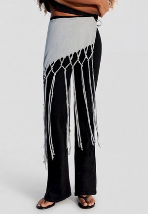 Étole en tissu gris avec franges nouées et perles portée sur un pantalon noir évasé et des sandales noires sur une personne debout.
