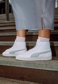 Vita hög-top sneakers har en ljusgrå sidostrand, texturerad gummisul och subtila varumärkesdetaljer. Bärs med rosa strumpor och en hundtandsmönstrad kjol.