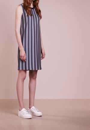 Junge Person trägt ein ärmelloses, vertikal schwarz-weiß gestreiftes Kleid mit weißen Sneakers und steht vor einem neutralen Hintergrund.