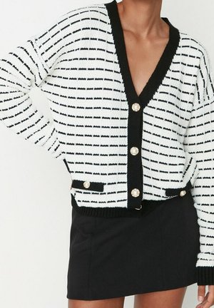 Frau trägt eine weiß-schwarz gestreifte Strickjacke mit Perlenknöpfen und einen schwarzen Minirock, Hand in die Hüfte gestützt.